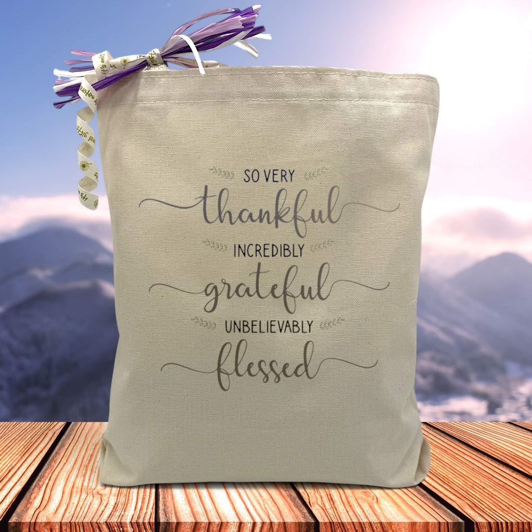  a grey stacked text saying so very thankful, incredibly grateful, unbelievably blessed printed on a canvas tote bag set on a wooden table with a mountain filled background,