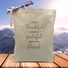  a grey stacked text saying so very thankful, incredibly grateful, unbelievably blessed printed on a canvas tote bag set on a wooden table with a mountain filled background,