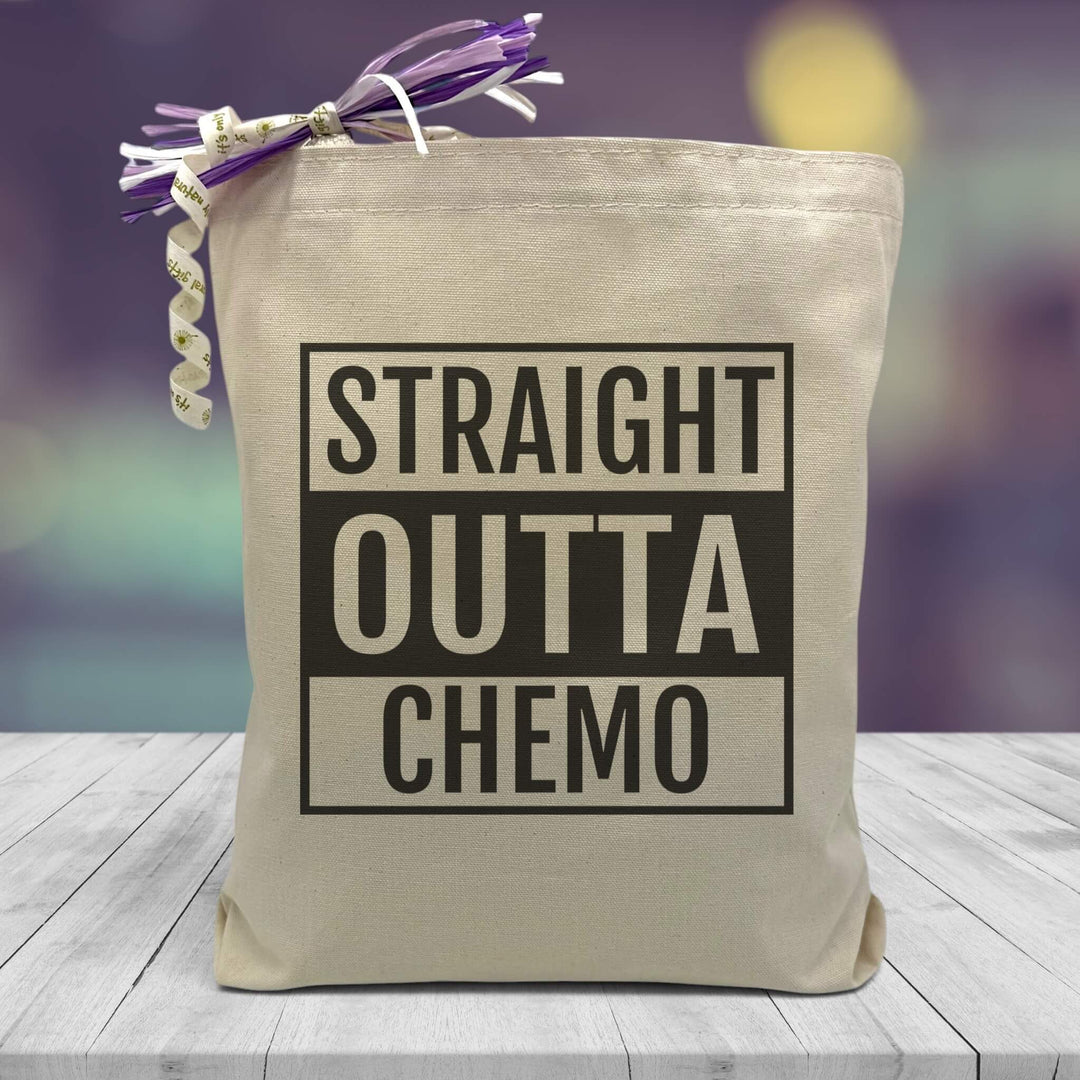 a text saying straight outta chemo printed on a canvas tote bag set on a wooden table with a purple blurred background.