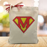 The super man logo with the letter m in the middle for a super mom theme printed on a canvas tote bag set on a wood table with a light filled background
