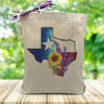  a red and blue water colored texas state with a star, sunflower and lavender in the middle printed on a canvas tote bag set on wooden table.