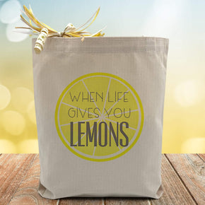 a lemon slice with a black text in the middle saying when life gives you lemons printed on a canvas tote bag.