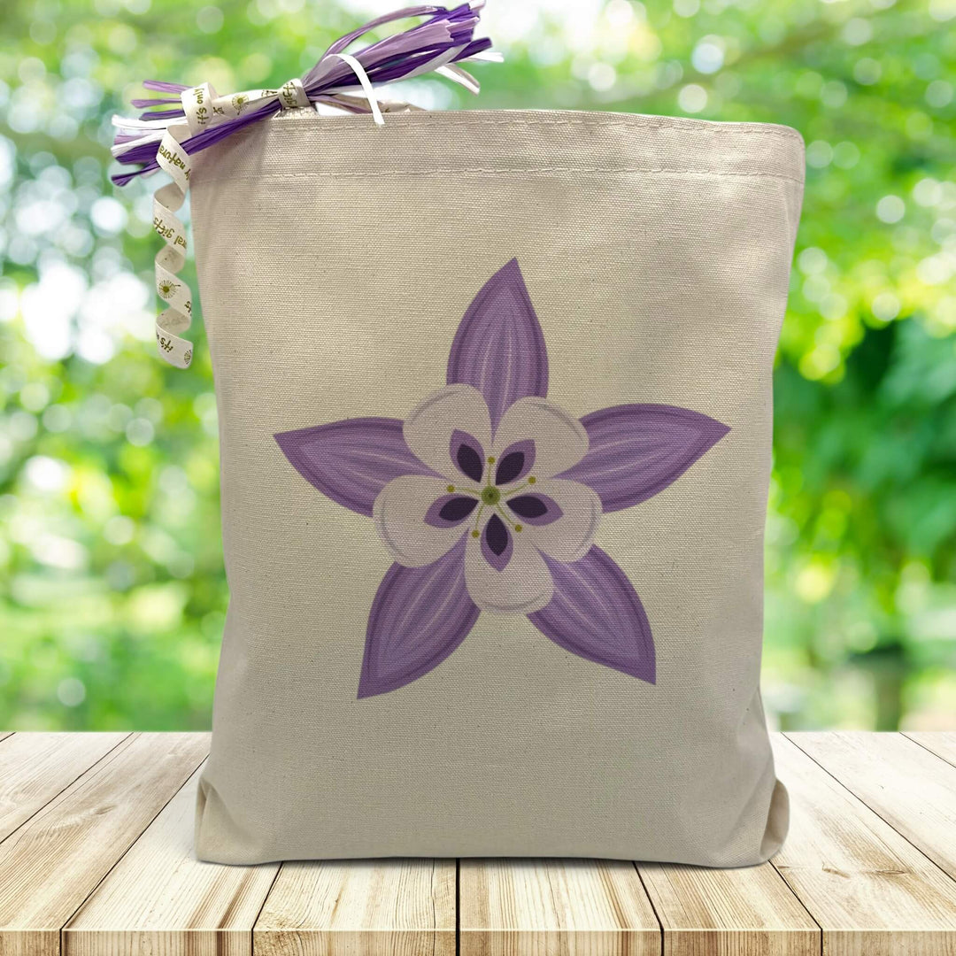 a purple columbine printed on a canvas tote bag set on a wooden table with a leafy background. 