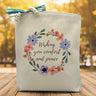a black text saying wishing you comfort and peace surrounded by a floral background printed on a canvas tote bag 