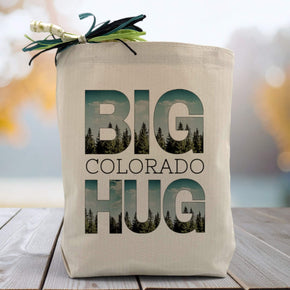 Big Colorado Hug text with tree line in background of text on a canvas tote bag sitting on wood table with blurred blue background.