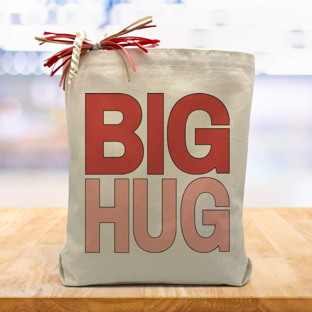 A pink and red big hug text on a canvas tote bag, placed on a wood table with a blurred blue background.