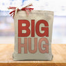 A pink and red big hug text on a canvas tote bag, placed on a wood table with a blurred blue background.
