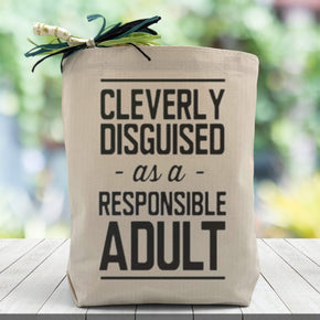 A all black cleverly disguised as a responsible adult text on a canvas tote bag placed on a wooden table with blurred green background.