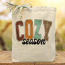 Cozy season printed on a canvas tote bag sitting on a table with a blurred background