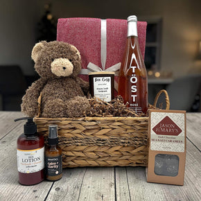 wicker basket with a blanket, teddy bear, candle, sparkling juice, lotion calming spritz, and caramels