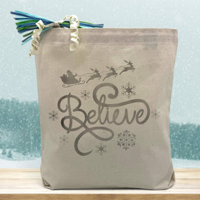 silver believe printed on a canvas tote bag sitting on a wood table with a snowy background