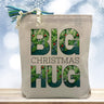 Big christmas hug with a pine tree theme printed on a canvas tote bag sitting on a wood table
