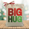 red and green big hug printed on a canvas tote bag sitting on a table with a christmas tree background