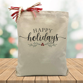 happy holidays printed on a canvas tote bag sitting on a wood table with a holiday blurred background