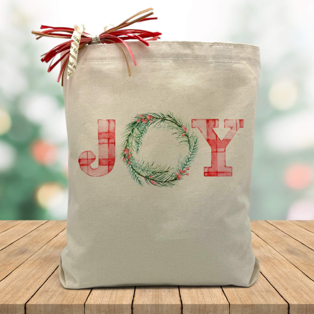 Joy printed on a canvas tote bag sitting on a wood table with a blurred holiday background