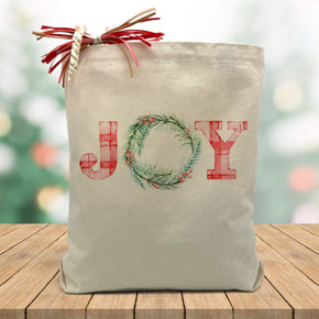 Joy printed on a canvas tote bag sitting on a wood table with a blurred holiday background