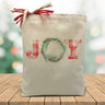 Joy printed on a canvas tote bag sitting on a wood table with a blurred holiday background