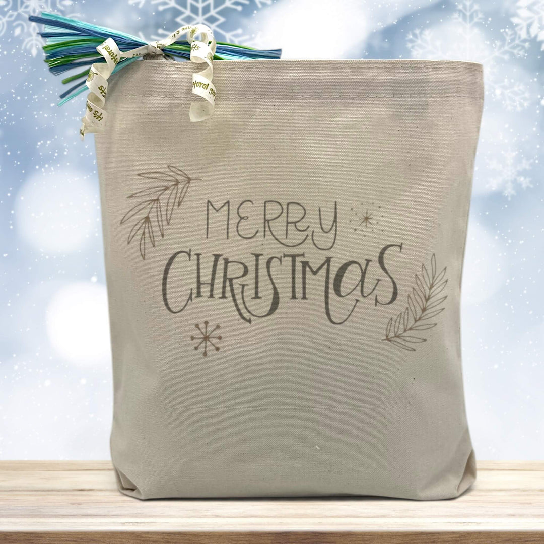 merry christmas printed on a canvas tote bag sitting on a wood table with a snowflake background
