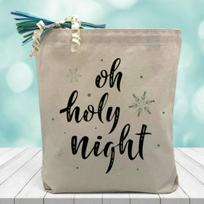 oh holy night printed on a canvas tote bag sitting on a wood table with a blurred blue background