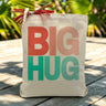 Big Hug in Pink/red and teal printed on a canvas tote bag sitting on an outdoor table
