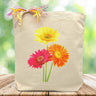 three daisies, yellow, pink and orange printed on a tote bag sitting on a wooden table with a green background