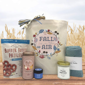 Gourmet fall gift basket with pretzels, tea, honey, a large candle and socks sitting on a table with a wheat field background