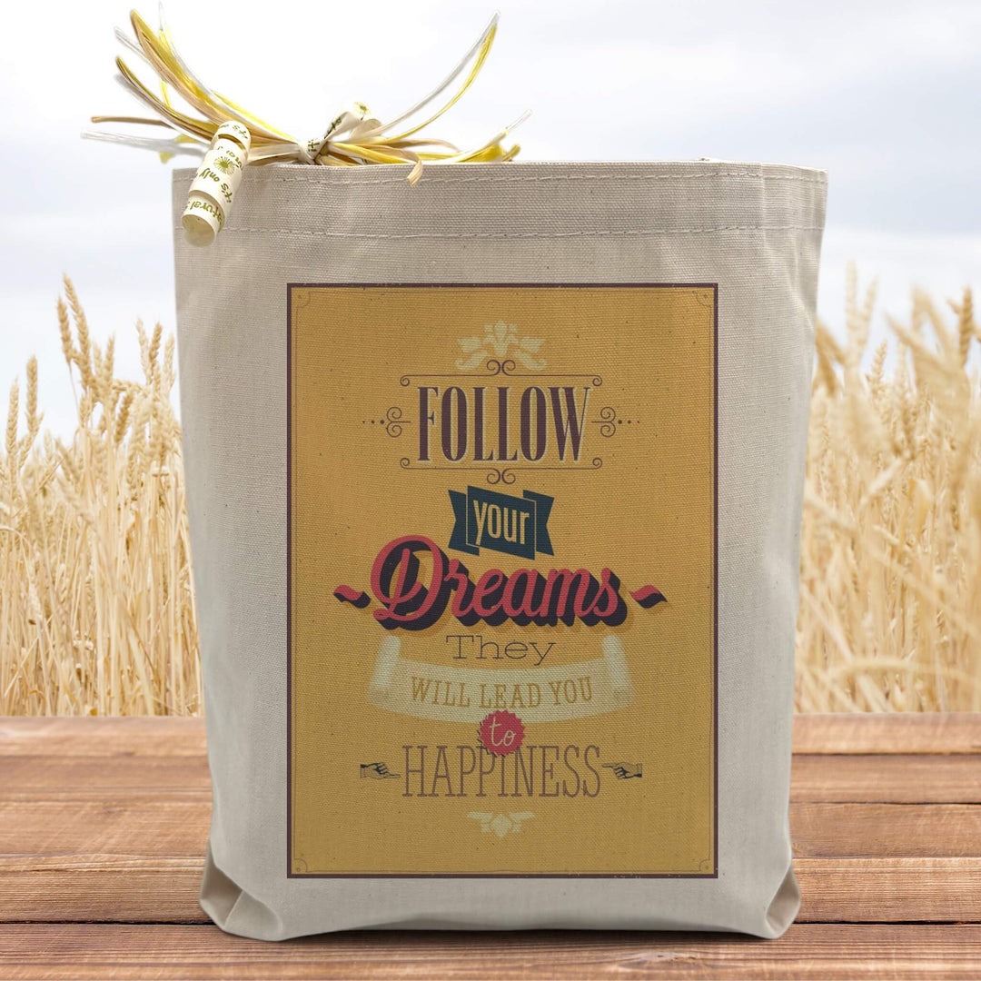 A yellow text filled box saying follow your dreams they will lead you to happiness on a canvas tote bag, placed on a table with a field in background. 