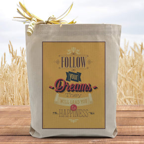 A yellow text filled box saying follow your dreams they will lead you to happiness on a canvas tote bag, placed on a table with a field in background. 