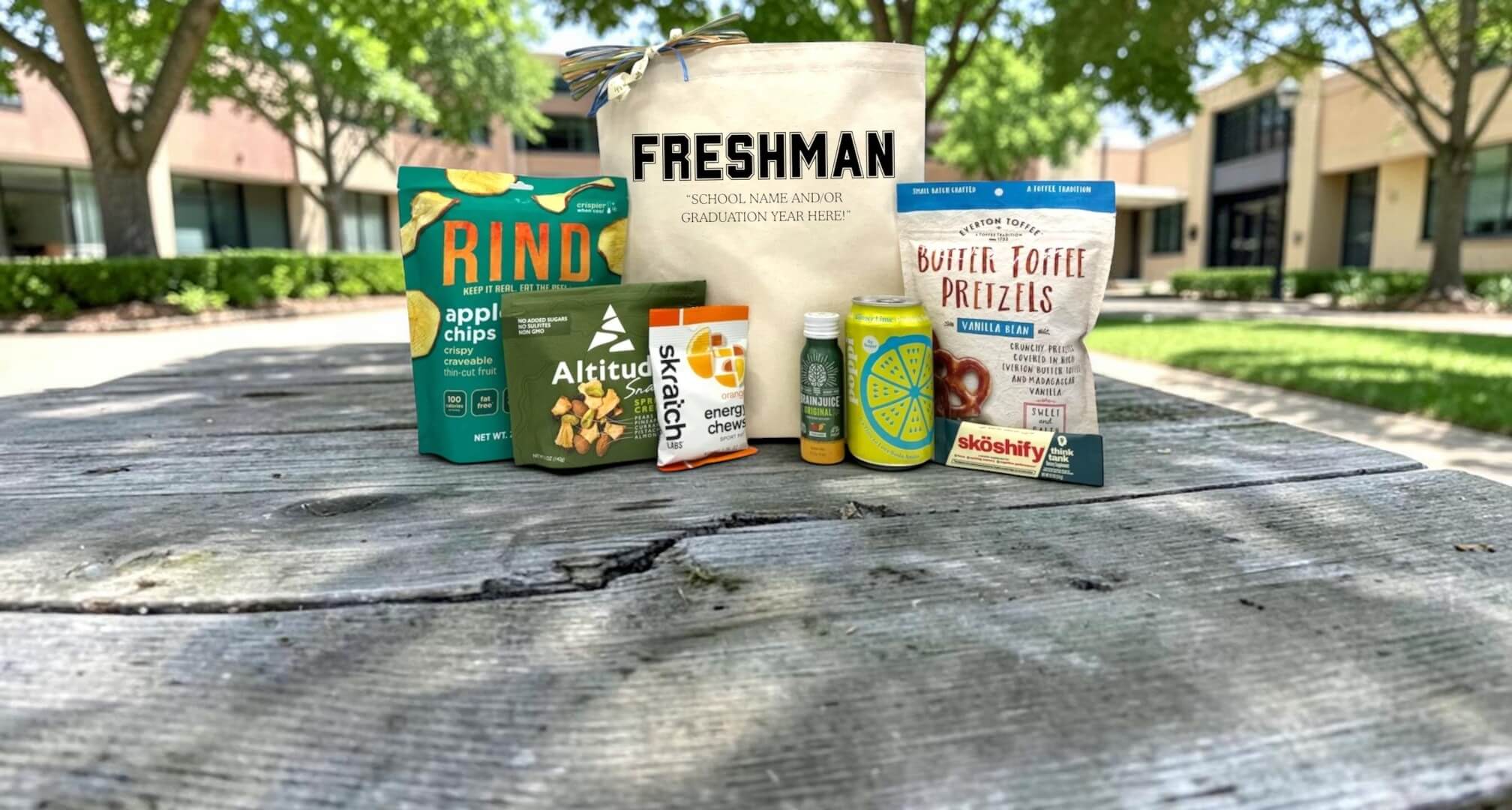 a freshman gift basket with snacks and evergy style foods sitting on a picnic table with a school in the background