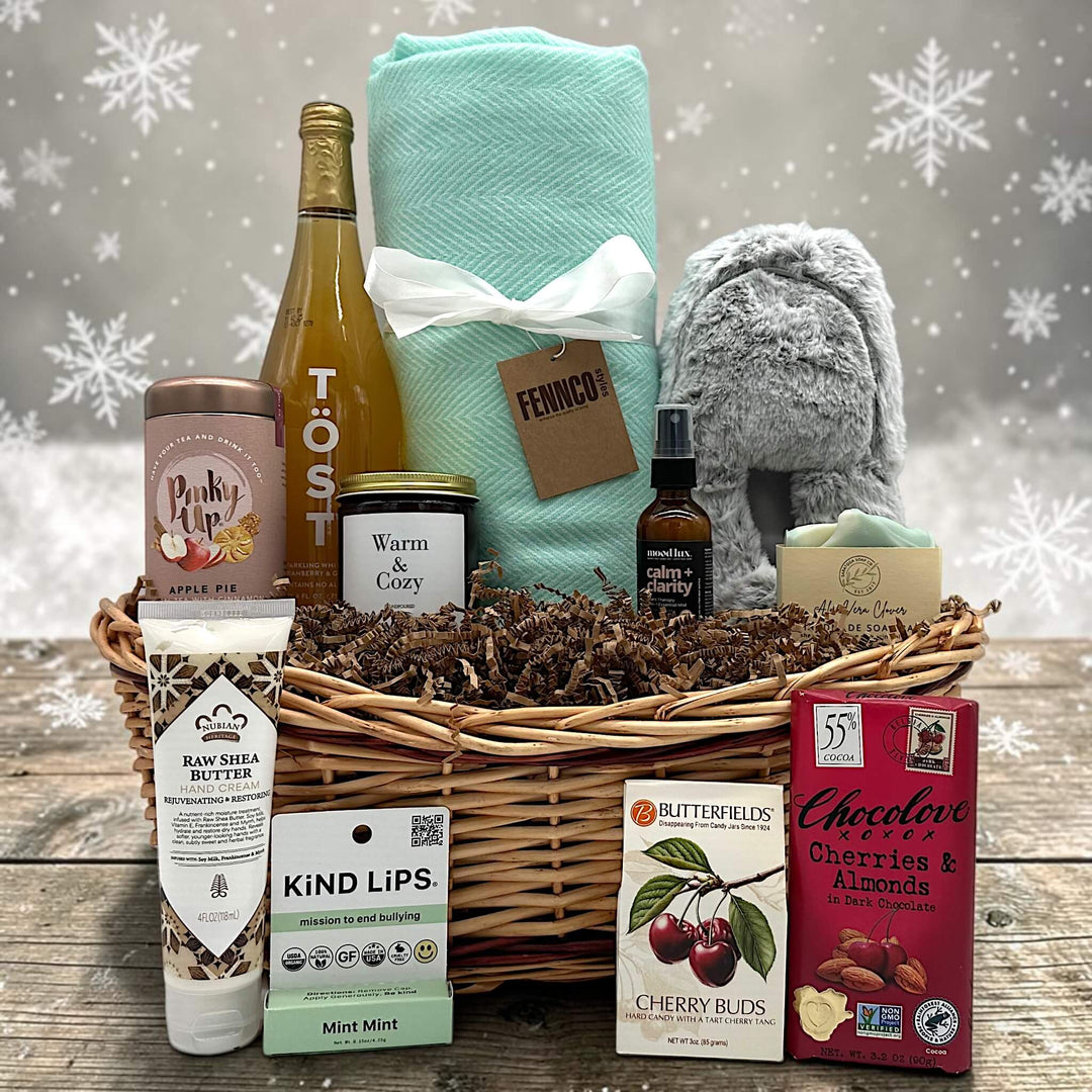 Gourmet cozy holiday gift basket with sparkling juice, a blanket, slippers, tea, a large candle, soap, lotion, and more sweets