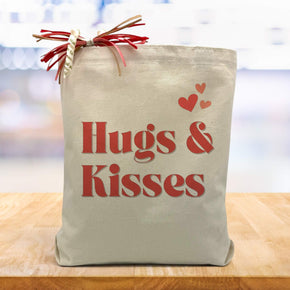 hugs & kisses with red hearts printed on a canvas tote bag sitting on a wood table with a lburred white and purple background