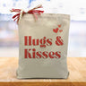 hugs & kisses with red hearts printed on a canvas tote bag sitting on a wood table with a lburred white and purple background