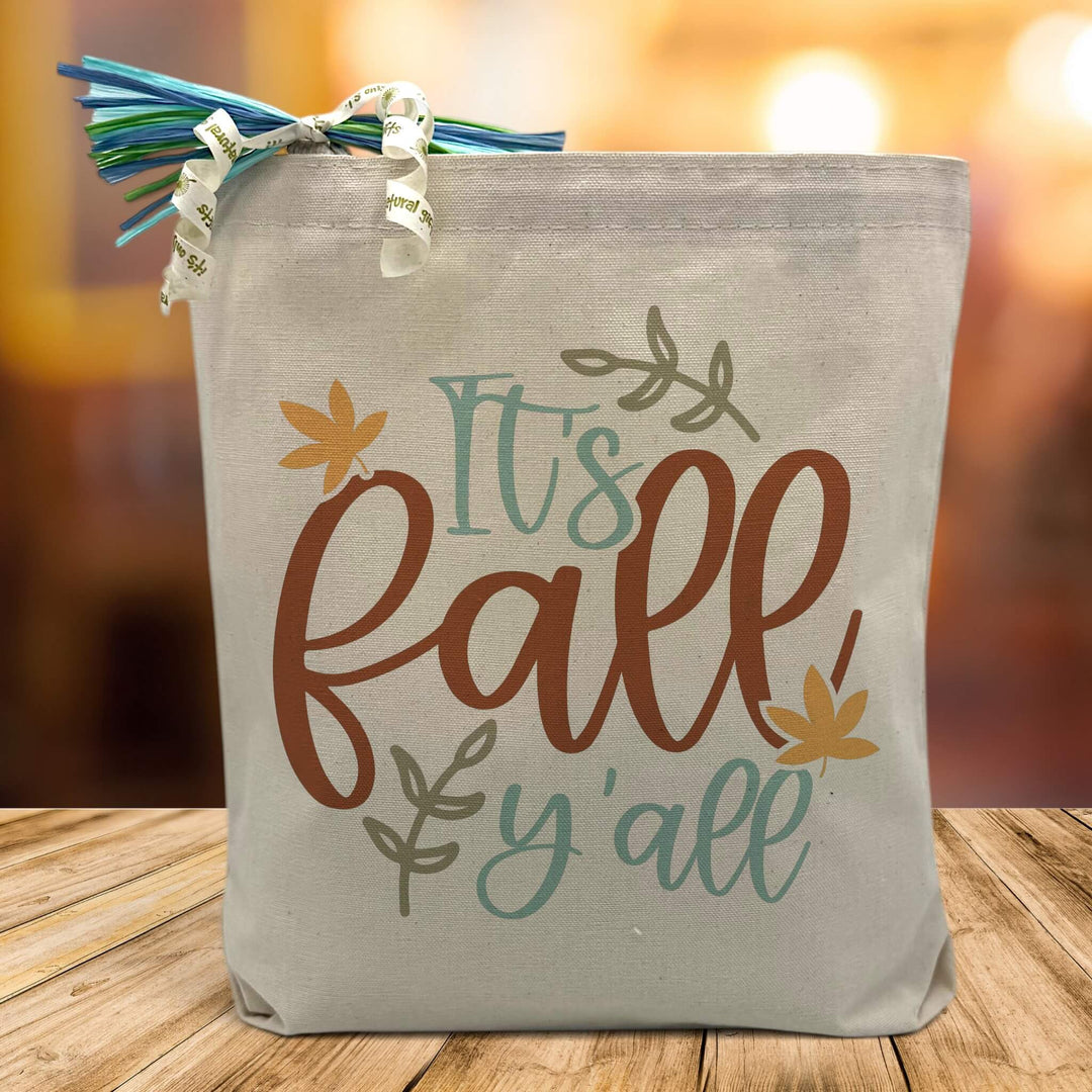 it's fall y'all designed printed on a canvas tote bag sitting on a table with a blurred fall background