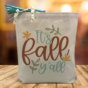 it's fall y'all designed printed on a canvas tote bag sitting on a table with a blurred fall background