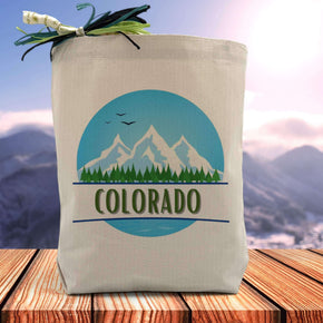 a mountain, trees and bird filled background with the word colorado placed in the middle printed on a canvas tote bag set on a wooden table 
