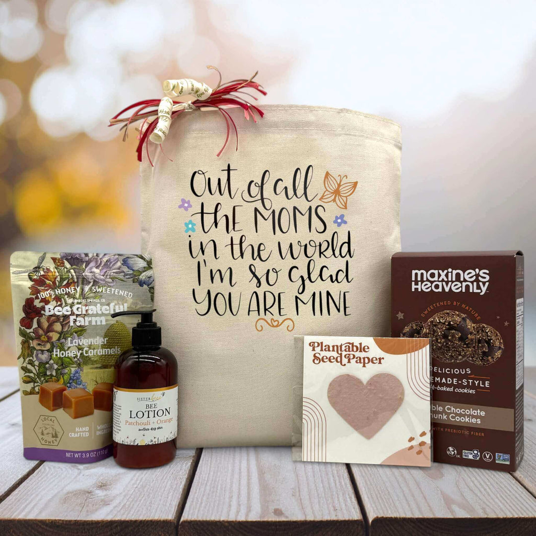 Quote out of all the moms in the world im so glad you are mine printed on a canvas totate bag with caramels, lotion, seed paper and cookies sitting on a table