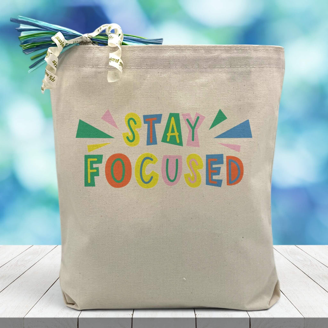 colorful stay focued printed on canvas tote bag sitting on table with blue blurred background