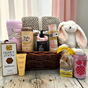 wicker basket with blanket, swaddle, tea, shampoo, teething ring, dishtowel, bunny, honey, lotion, caramels, and poppi