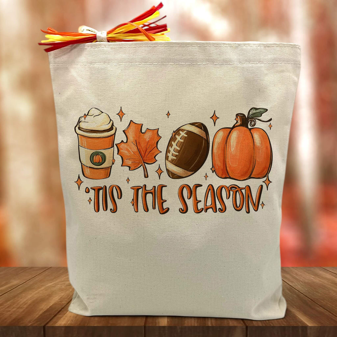 coffee, fall leaf, football and pumpkin with tis the season printed on a canvas tote bag sitting on a table